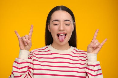 Photo of Happy woman showing her tongue and rock gesture on orange background