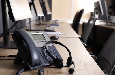 Photo of Stationary phone and headset near modern computer on wooden desk in office. Hotline service