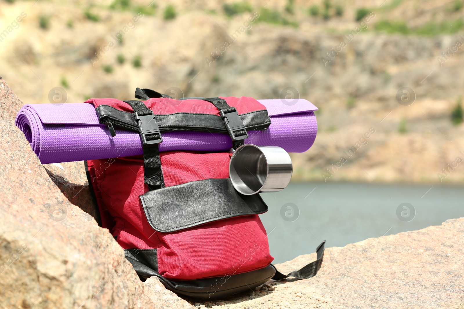 Photo of Camping backpack with mat and mug on stones near river. Space for text