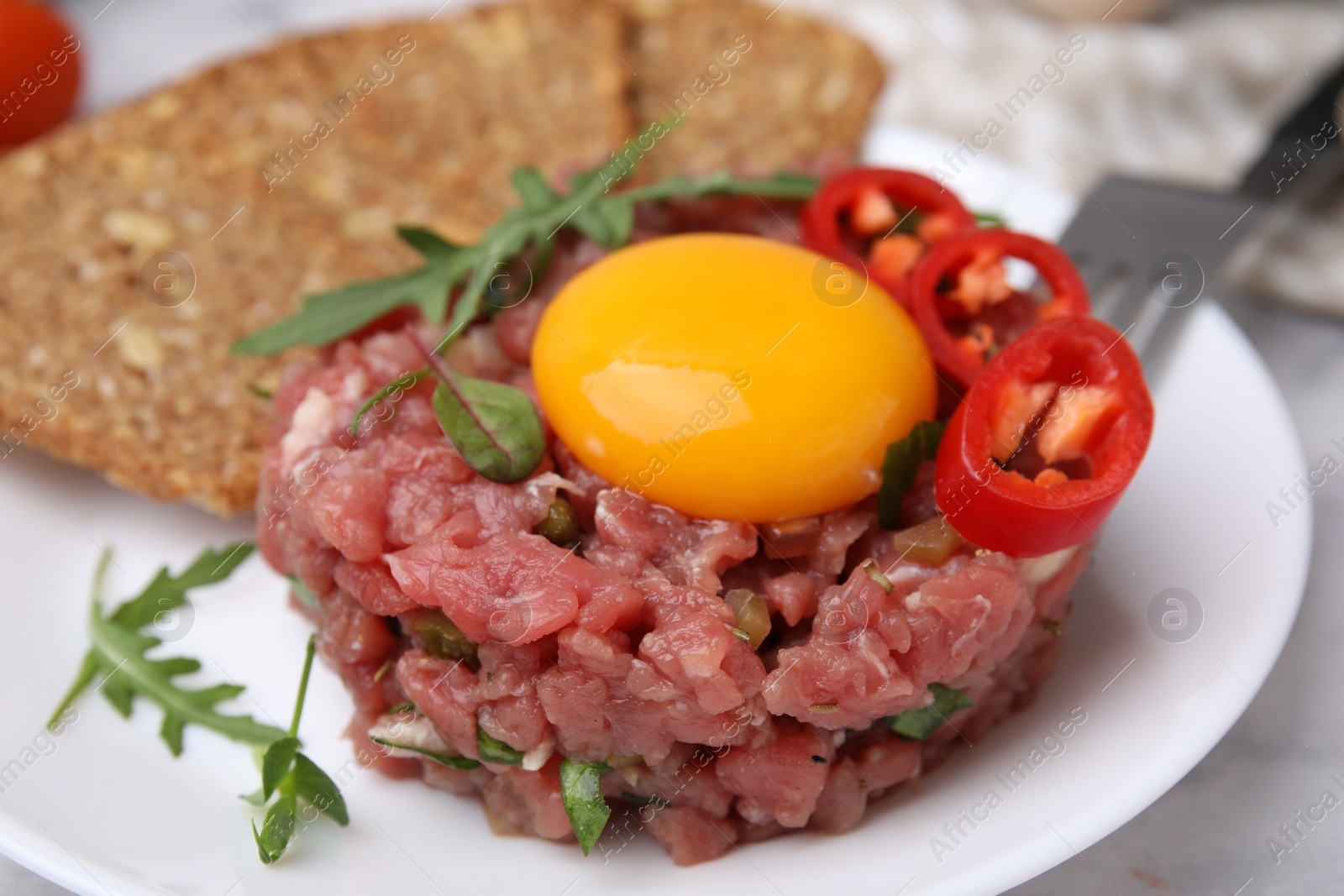 Photo of Tasty beef steak tartare served with yolk, pepper, bread and greens on white table, closeup