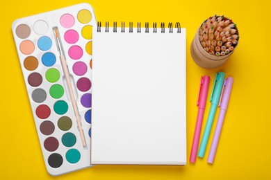 Watercolor palette, empty notebook, colorful pencils and markers on yellow background, flat lay. Space for text