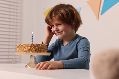 Cute boy with birthday cake at white table indoors