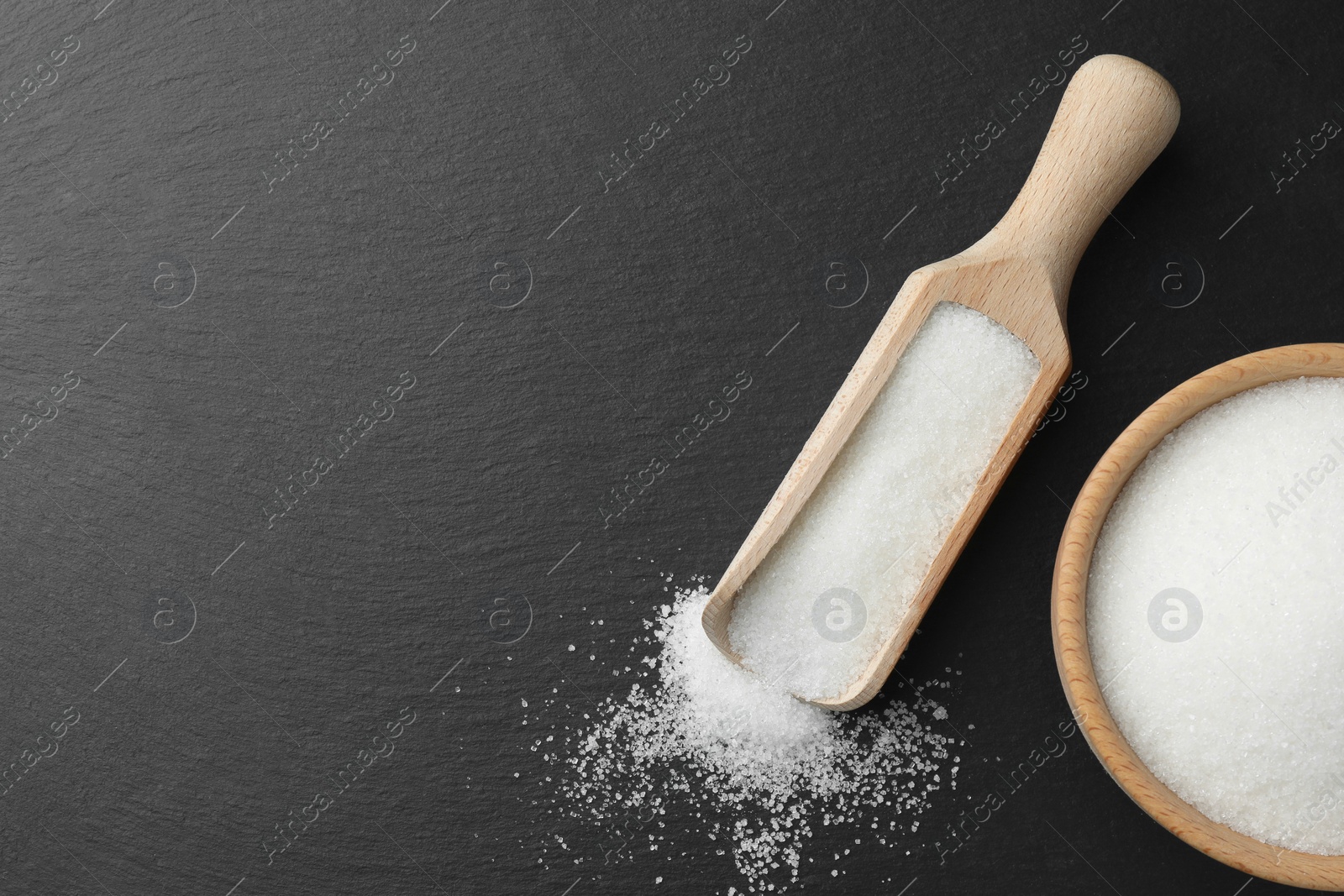 Photo of Granulated sugar in bowl and scoop on black table, flat lay. Space for text