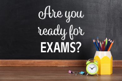 Image of Pencils and alarm clock on wooden table near blackboard with phrase ARE YOU READY FOR EXAMS