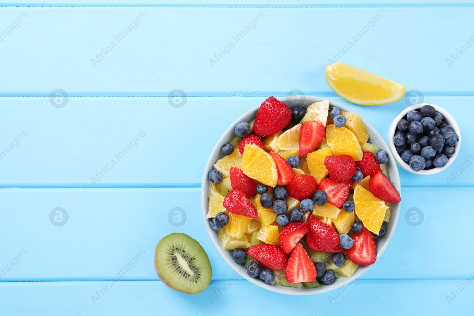 Photo of Delicious fresh fruit salad in bowl and ingredients on light blue wooden table, flat lay. Space for text