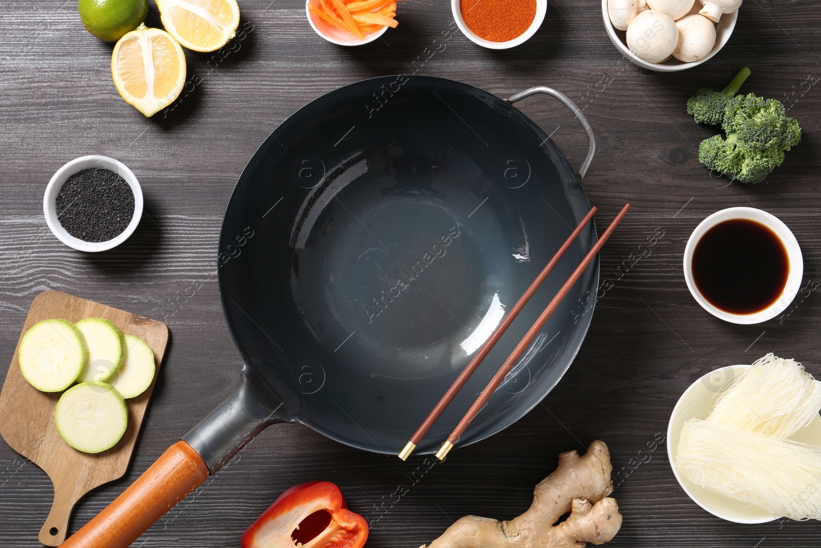 Photo of Empty iron wok and chopsticks surrounded by ingredients on dark grey wooden table, flat lay