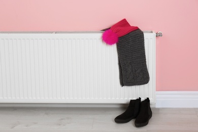 Photo of Heating radiator with knitted cap, scarf and shoes near color wall