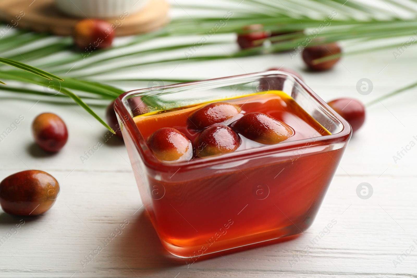 Photo of Palm oil in glass bowl with fruits and tropical leaves on white wooden table