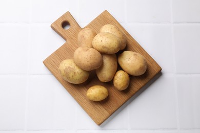 Photo of Raw fresh potatoes and wooden board on white tiled table, top view