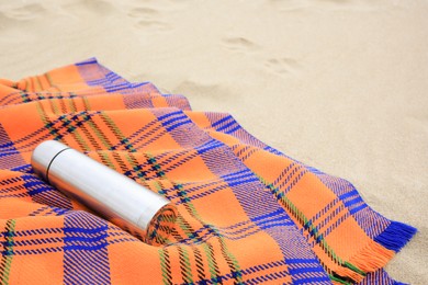 Metallic thermos with hot drink and plaid on sandy beach, space for text