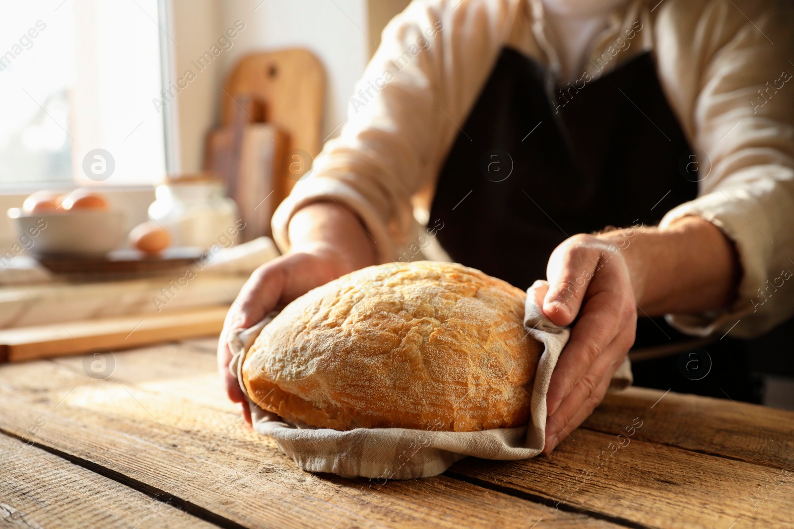 Photo of Man holding loaf of fresh bread at wooden table indoors, closeup