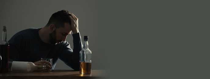 Image of Suffering from hangover. Man with alcoholic drink at table against grey background, space for text. Banner design