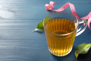 Photo of Cup of herbal diet tea, measuring tape and leaves on blue wooden table, space for text. Weight loss concept