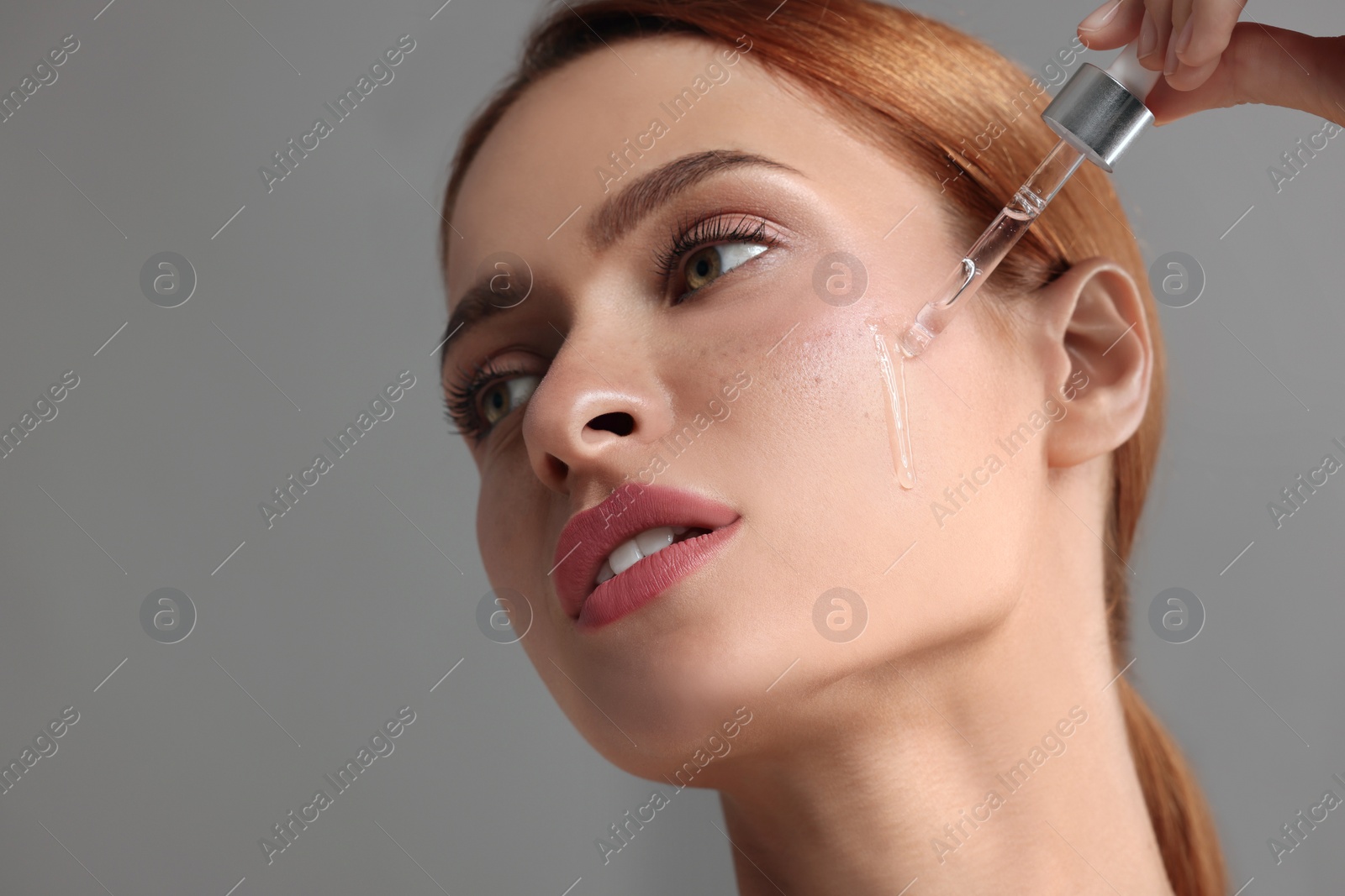 Photo of Beautiful young woman applying cosmetic serum onto her face on grey background, space for text