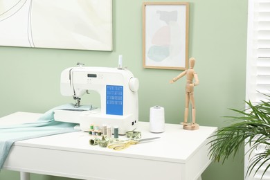 Modern sewing machine with cloth and craft accessories on white table near light green wall