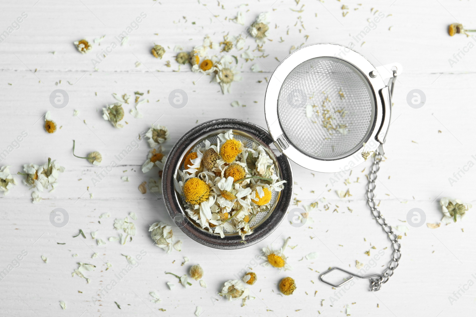 Photo of Dry chamomile flowers in infuser on white wooden table, flat lay