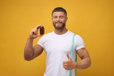 Athletic young man with measuring tape and bottle of supplements showing thumb up gesture on orange background. Weight loss