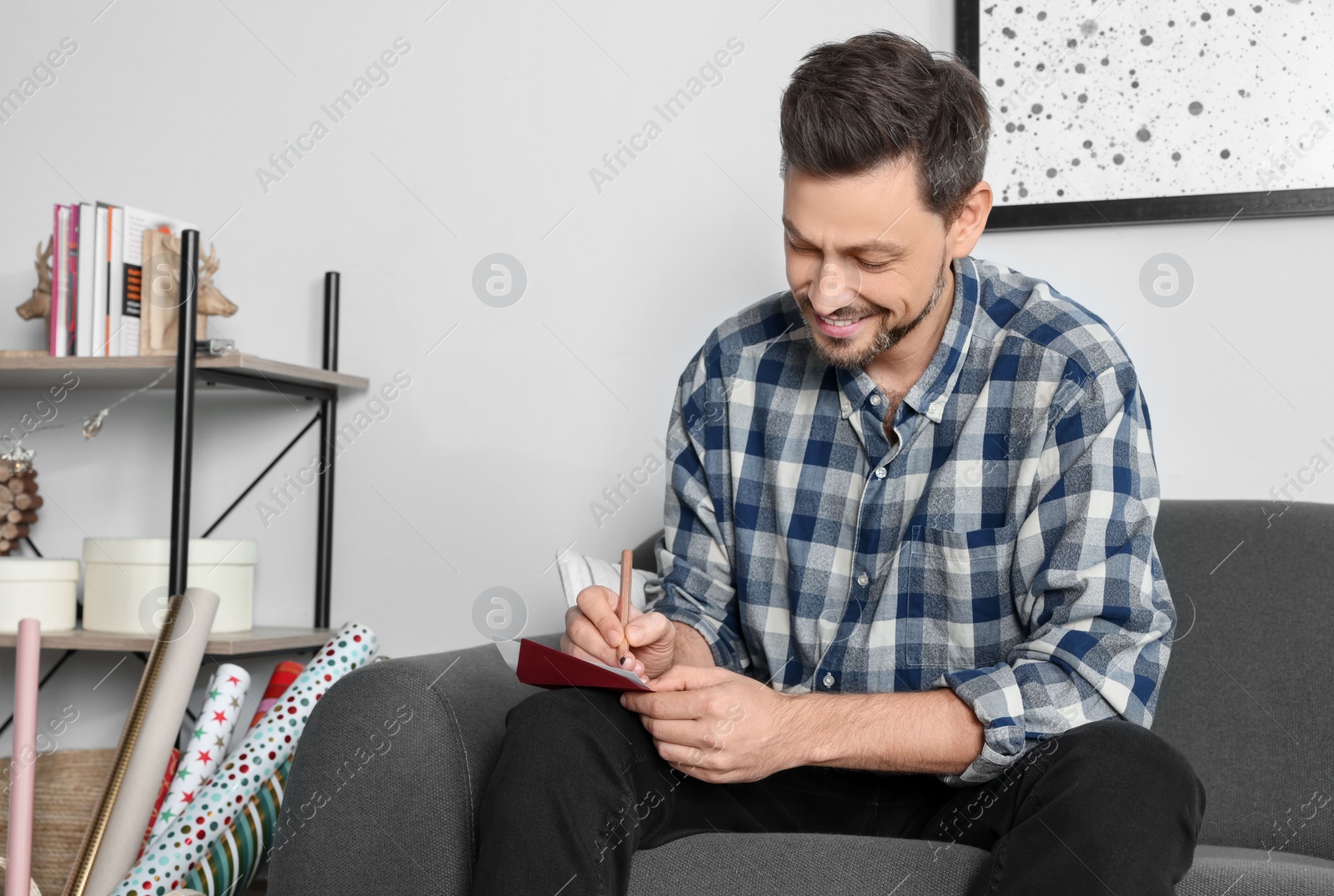 Photo of Happy man writing message in greeting card on sofa in living room