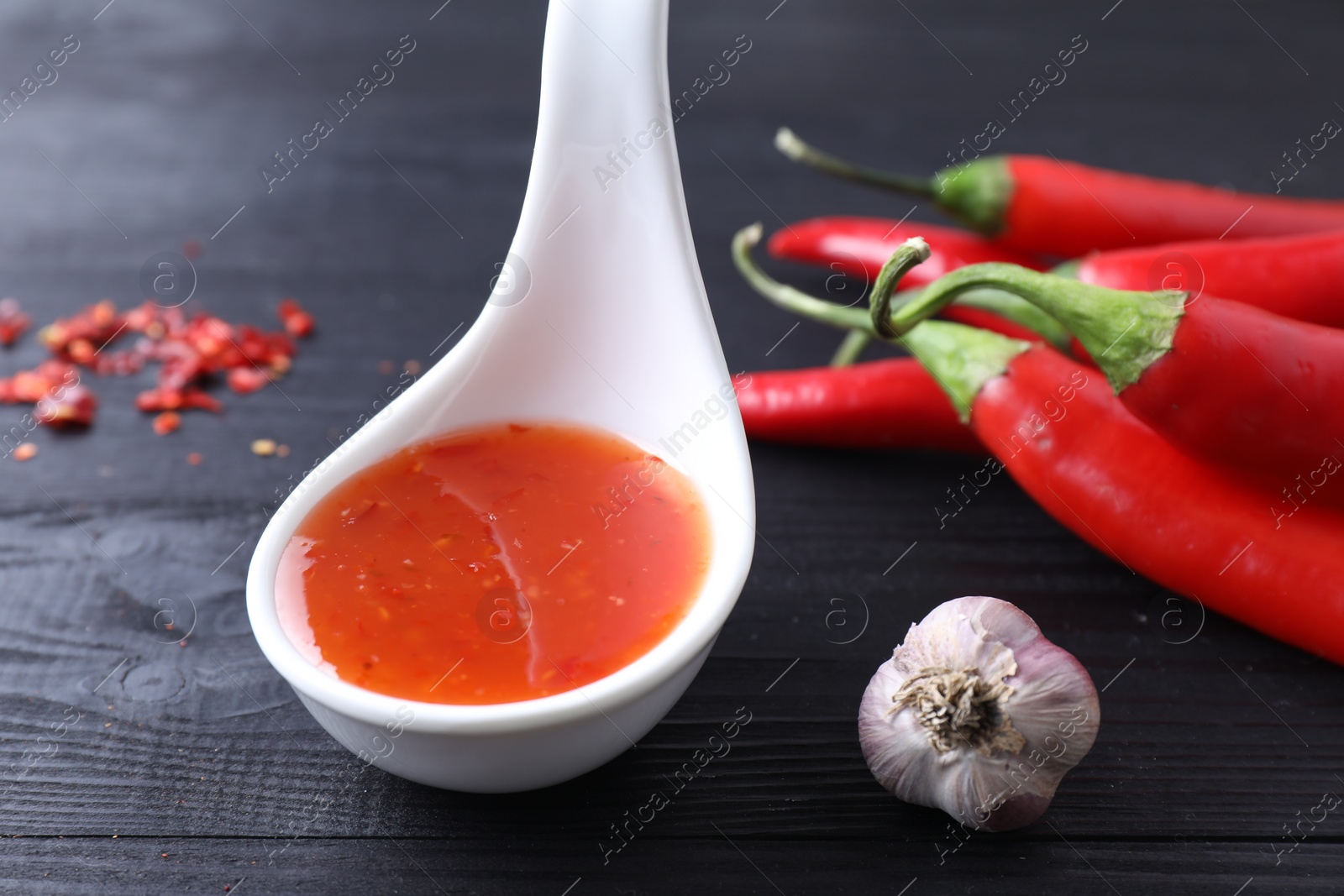 Photo of Spicy chili sauce in spoon, peppers and garlic on black wooden table, closeup