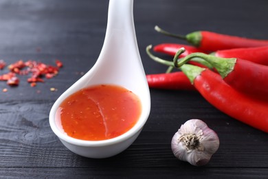 Photo of Spicy chili sauce in spoon, peppers and garlic on black wooden table, closeup