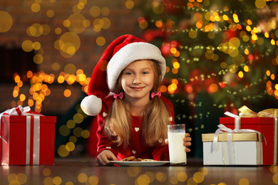 Photo of Little girl in Santa Claus cap with cookies and milk near Christmas tree indoors