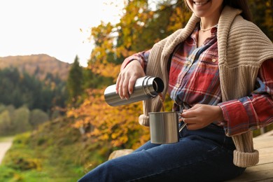 Photo of Woman pouring hot drink from thermos into metallic mug outdoors, closeup. Space for text