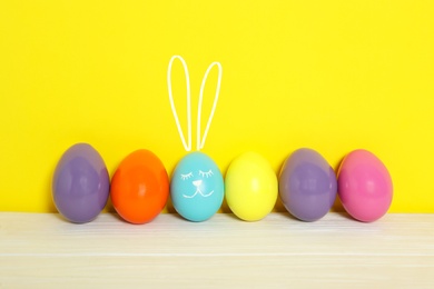 Image of One egg with drawn face and ears as Easter bunny among others on white wooden table against yellow background