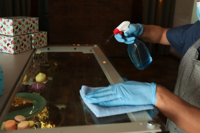 Photo of Worker in gloves disinfecting dessert showcase at cafe, closeup