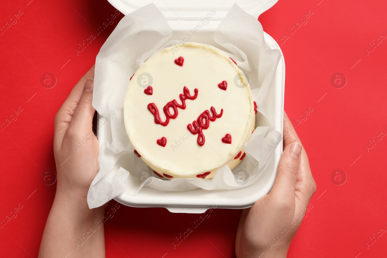 Photo of Woman holding takeaway box with bento cake at red table, top view. St. Valentine's day surprise