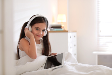 Photo of Cute little girl with headphones and tablet listening to audiobook in bed at home