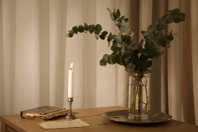 Photo of Beautiful eucalyptus branches, old book and holder with burning candle on wooden table indoors. Interior element