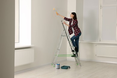 Photo of Young woman painting wall with roller on stepladder indoors. Room renovation