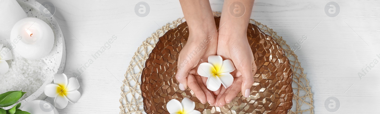 Image of Spa treatment. Woman soaking her hands in bowl with water and flowers on white wooden table, top view. Banner design