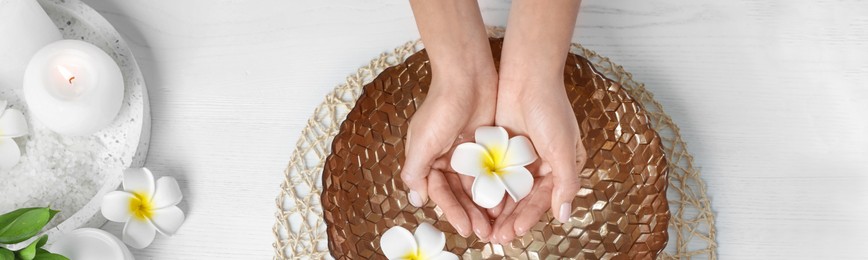 Image of Spa treatment. Woman soaking her hands in bowl with water and flowers on white wooden table, top view. Banner design