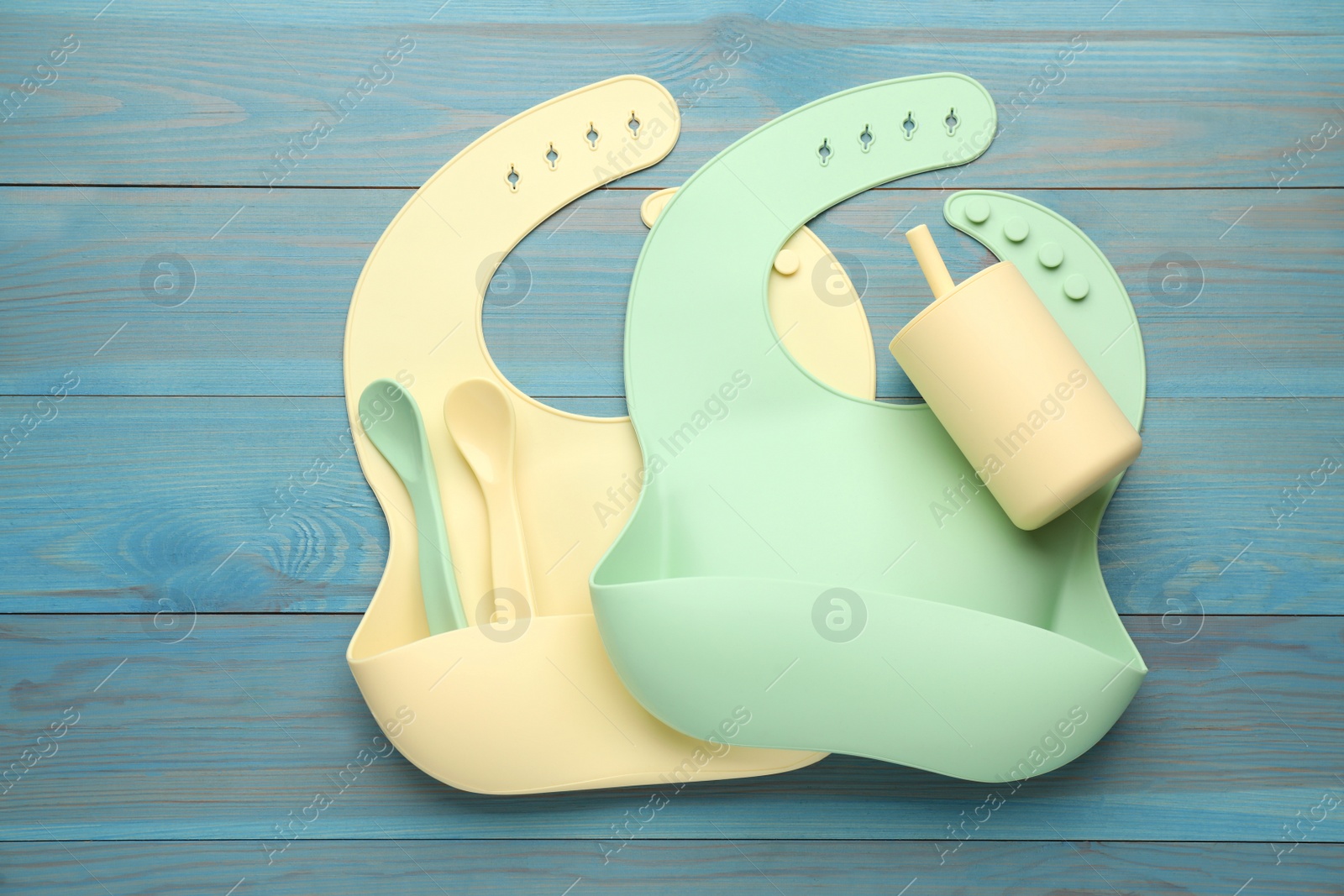 Photo of Flat lay composition with silicone baby bibs and plastic dishware on light blue wooden background