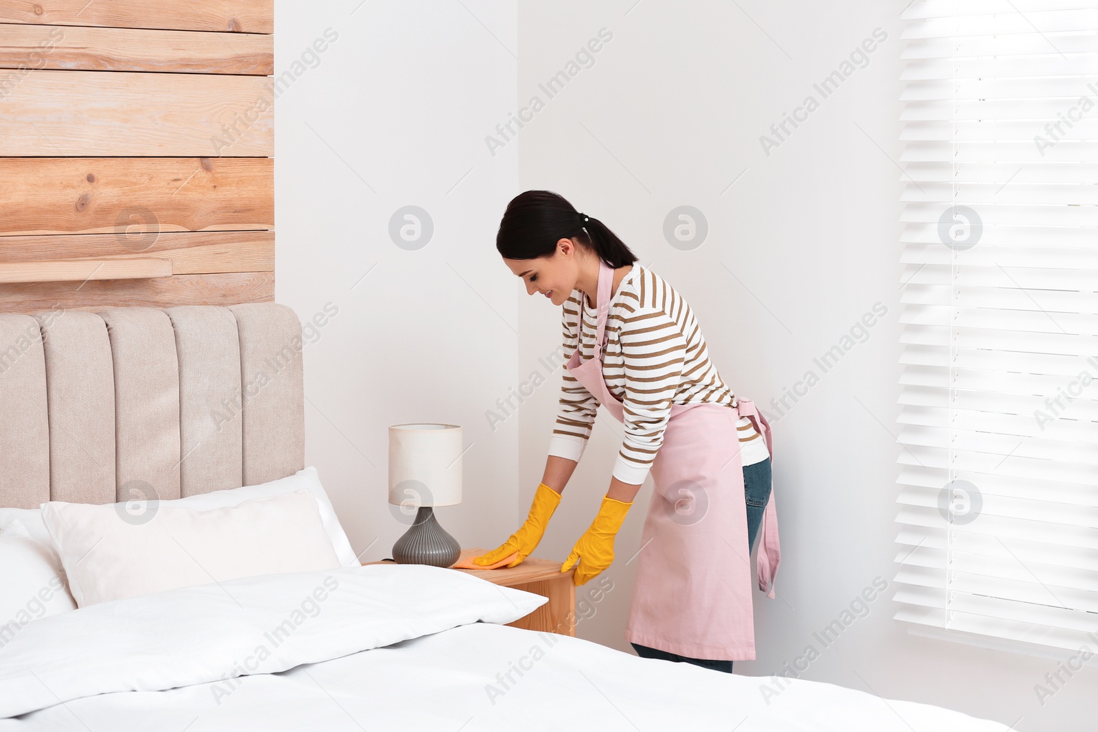 Photo of Young chambermaid wiping dust from nightstand in bedroom