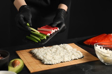 Photo of Chef in gloves putting cucumber onto unwrapped sushi roll at dark table, closeup