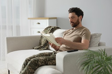 Photo of Soldier reading book on soft sofa in living room. Military service