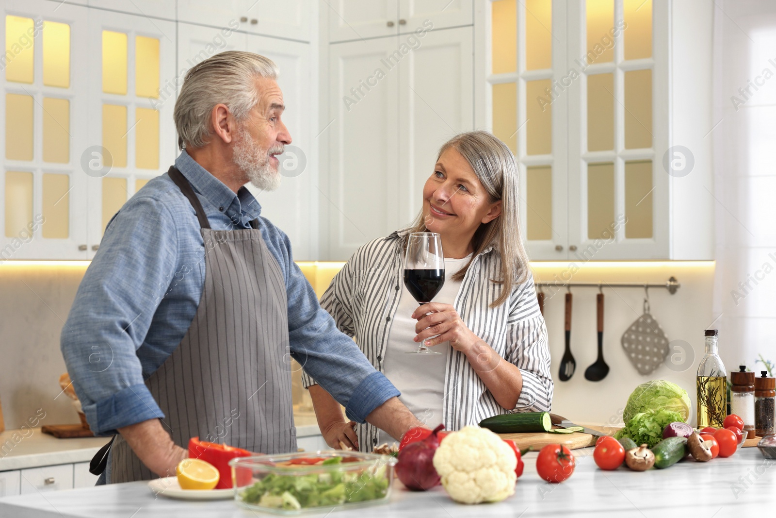 Photo of Happy senior couple cooking together in kitchen. Woman with glass of wine near her husband