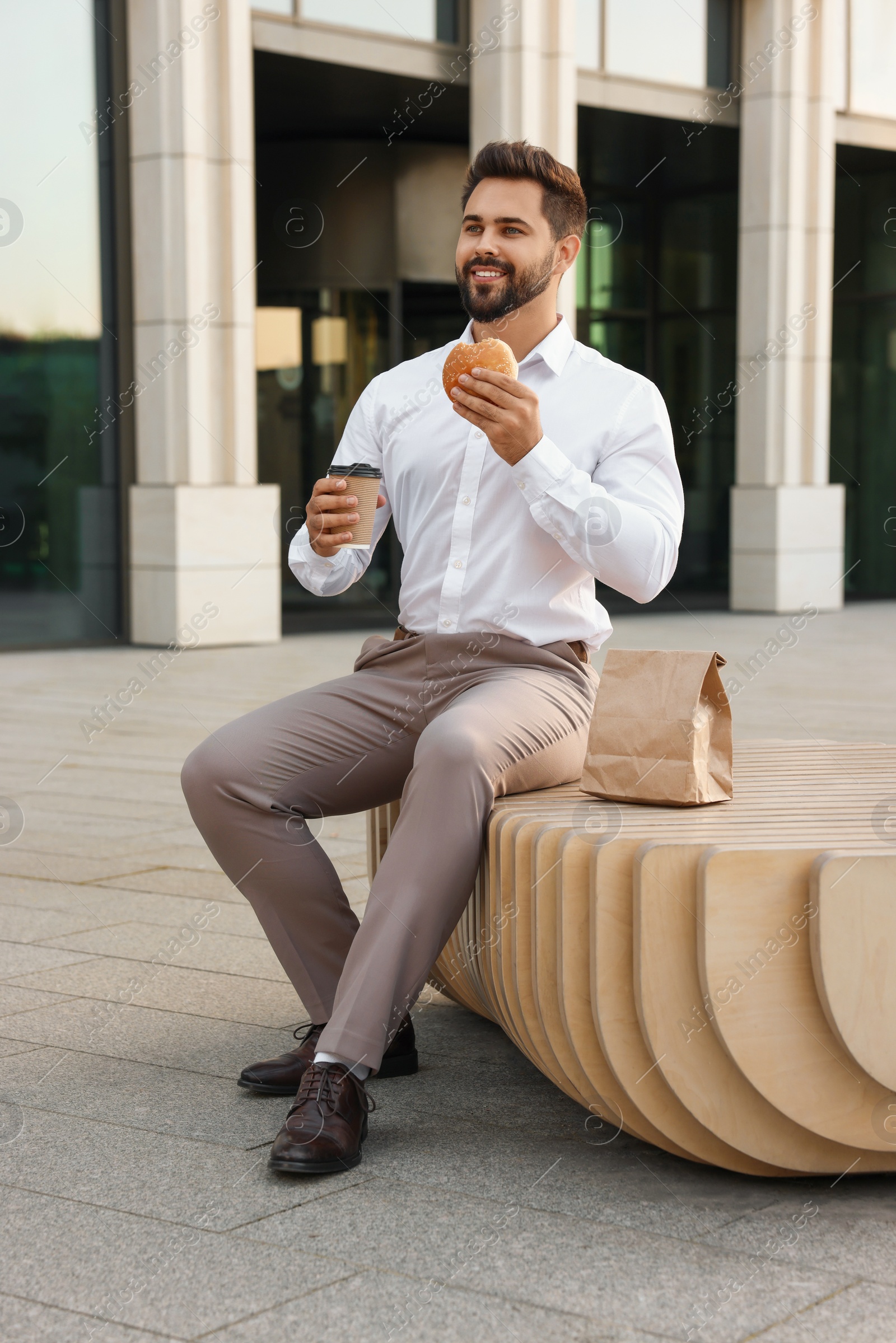 Photo of Businessman with hamburger and paper cup of coffee having lunch on bench outdoors