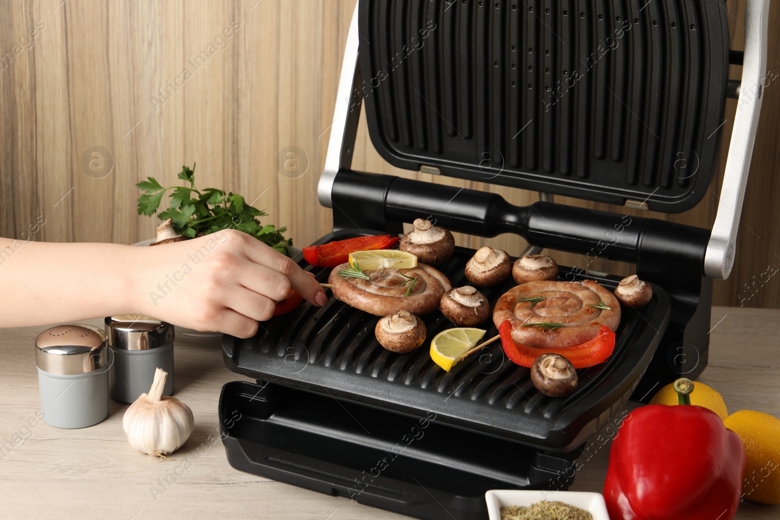 Photo of Woman cooking homemade sausages with mushrooms and bell pepper on electric grill at wooden table, closeup