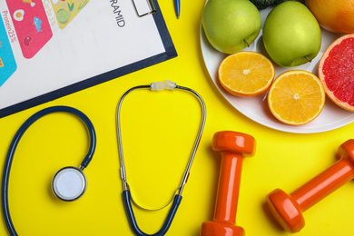 Fruits, dumbbells, stethoscope and list of products on yellow background, flat lay. Visiting nutritionist