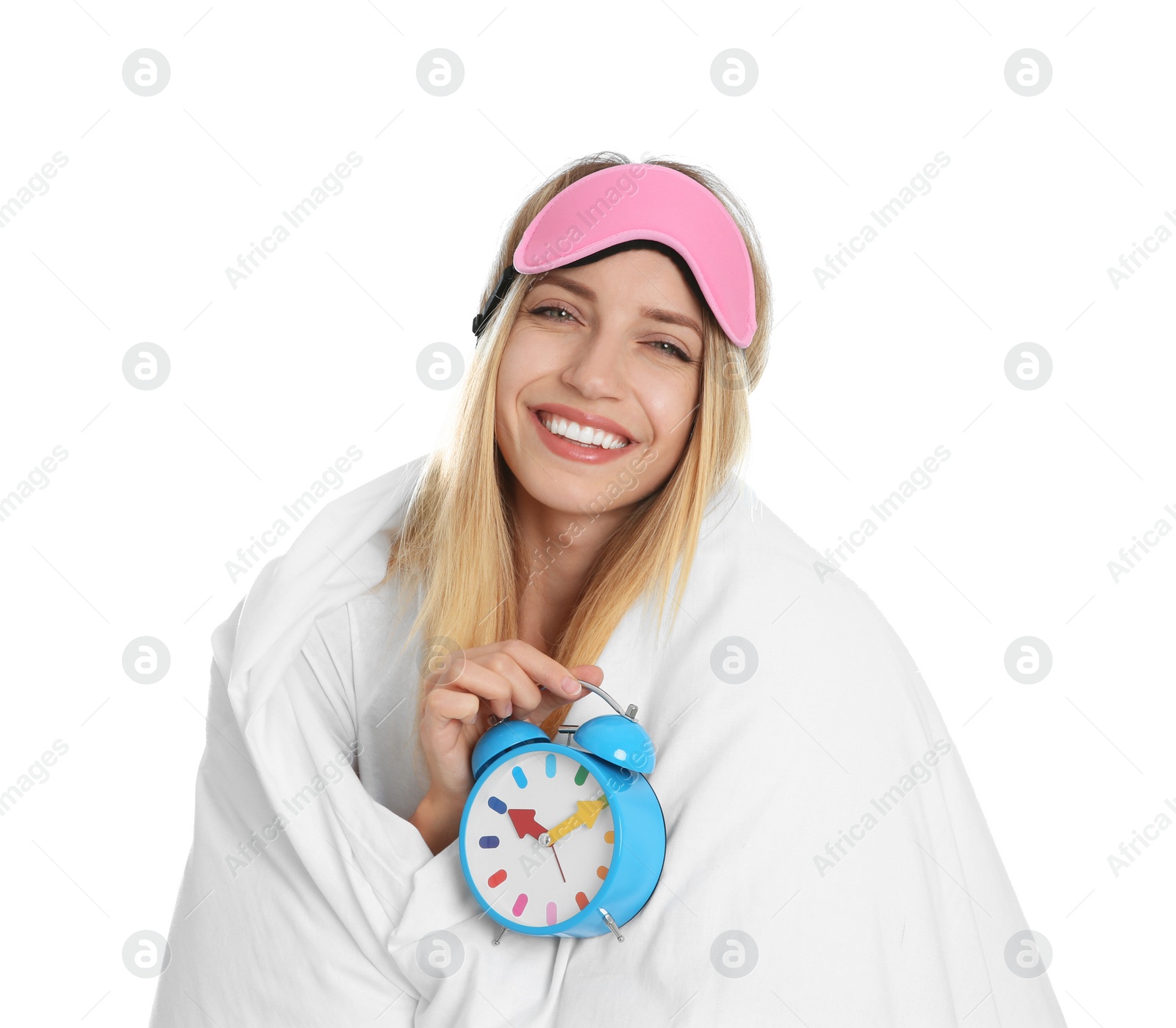 Photo of Young woman in sleeping mask wrapped with blanket holding alarm clock on white background