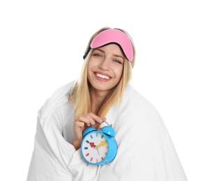 Photo of Young woman in sleeping mask wrapped with blanket holding alarm clock on white background