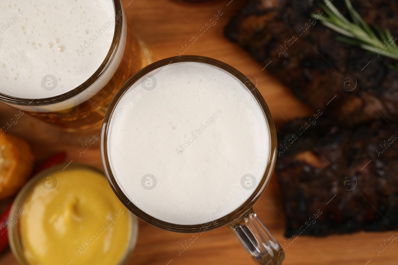 Photo of Mugs of beer, delicious grilled ribs and sauce on wooden table, closeup