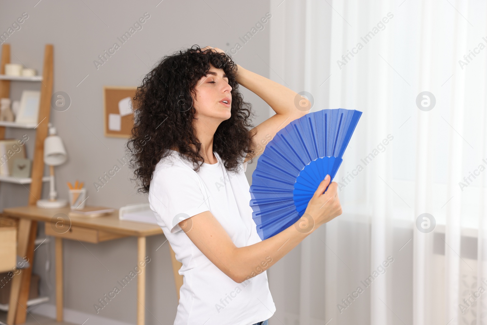 Photo of Young woman waving blue hand fan to cool herself at home