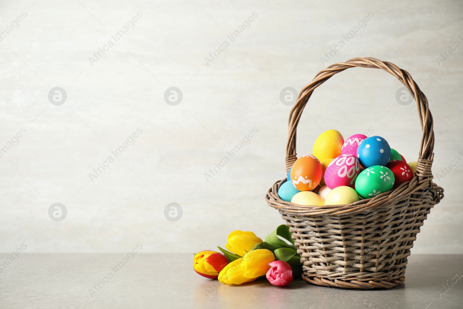 Photo of Basket with colorful Easter eggs and tulip flowers on light grey background. Space for text