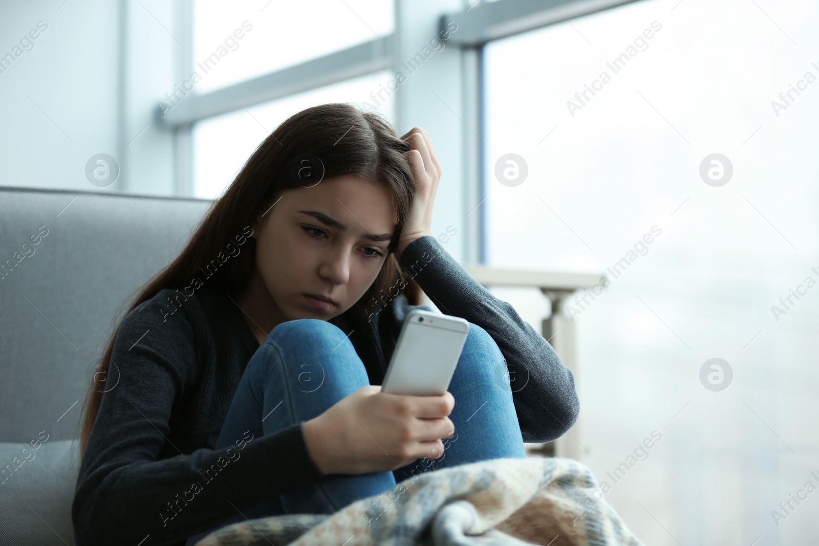Photo of Upset teenage girl with smartphone sitting at window indoors. Space for text