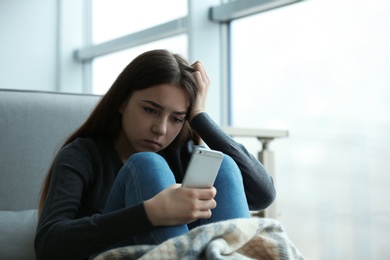 Upset teenage girl with smartphone sitting at window indoors. Space for text