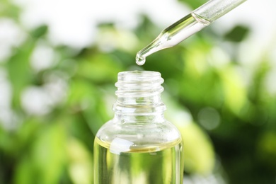 Dropper with essential oil over bottle against blurred background, closeup